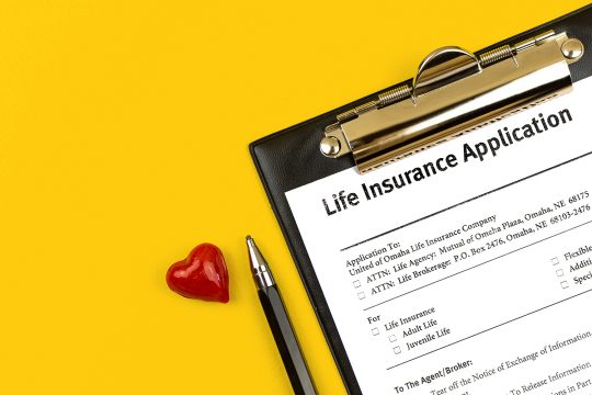 Life insurance application. Clipboard with agreement, pen and red heart on a yellow desktop. Top view